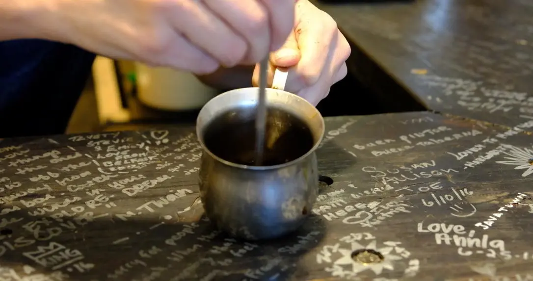 Cuban coffee being stirred in a small metal pot on a signed wooden counter