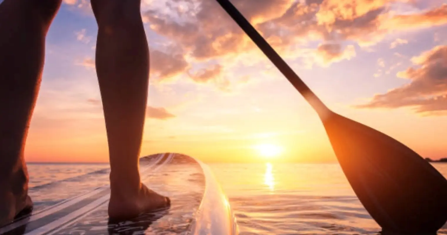 Feet on a paddle board with a paddle at sunset over the water