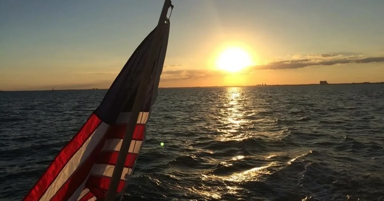 Sailboat mast, sail and an American flag silhouetted against the sunset